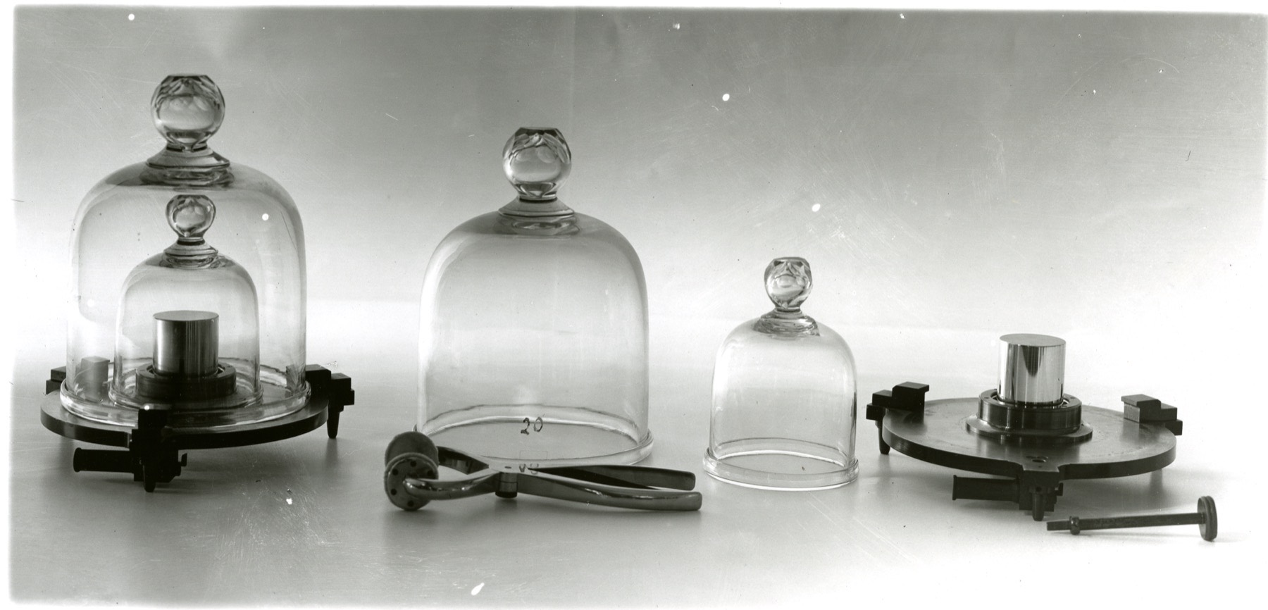 Mass standards under bell jars at the US National Bureau of Standards: Kilogram No. 20, the American primary standard, and Kilogram No. 4, with the tongs used to handle them. Photo: NIST, public domain.